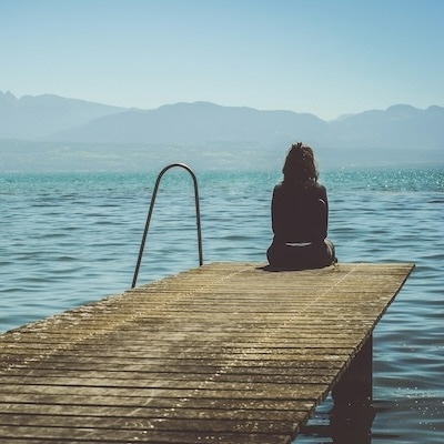 A woman sits on the end of a dock during daytime staring across a lake